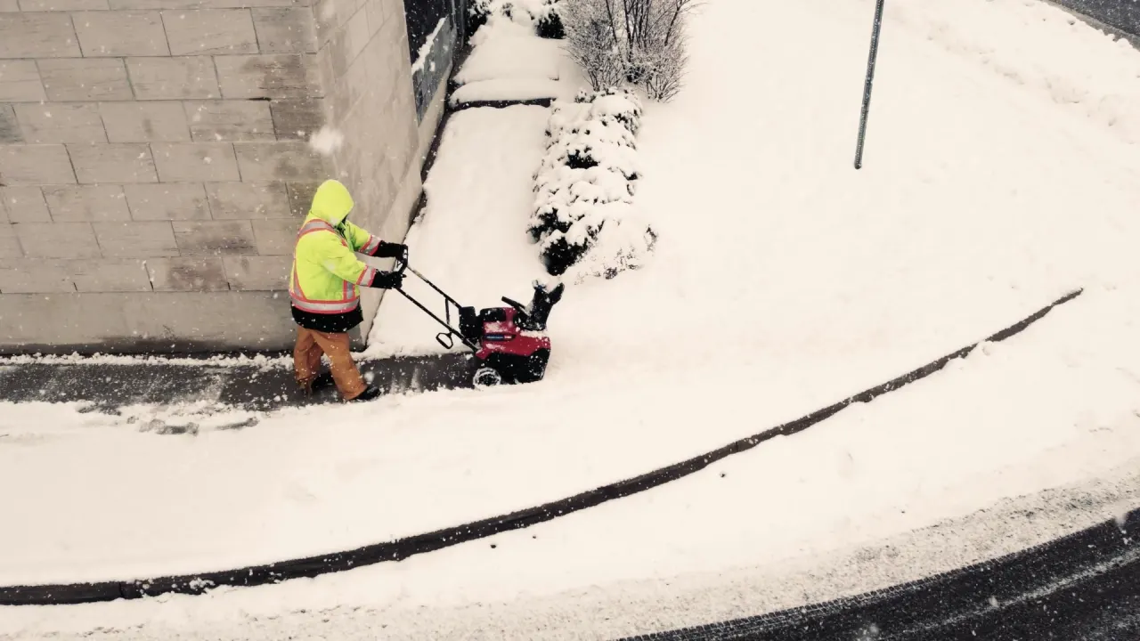 Worker in a safety jacket clearing a sidewalk with a snowblower
